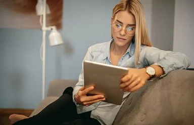 Eine Frau mit Brille sitzt auf einem Sofa und nutzt ein Tablet.