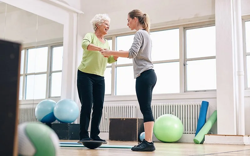 In einem Fitnesstudio trainiert eine ältere Frau auf einem Balance-Trainer ihr Gleichgewicht. Eine Trainerin steht vor ihr und hält sie fest.