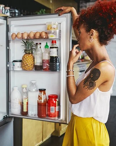 Eine Frau in einem sommerlichen Outfit steht überlegend vor dem gut gefüllten Kühlschrank, die geöffnetet Kühlschranktür in der Hand.