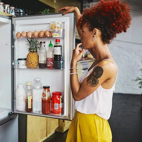 Eine Frau in einem sommerlichen Outfit steht überlegend vor dem gut gefüllten Kühlschrank, die geöffnetet Kühlschranktür in der Hand.
