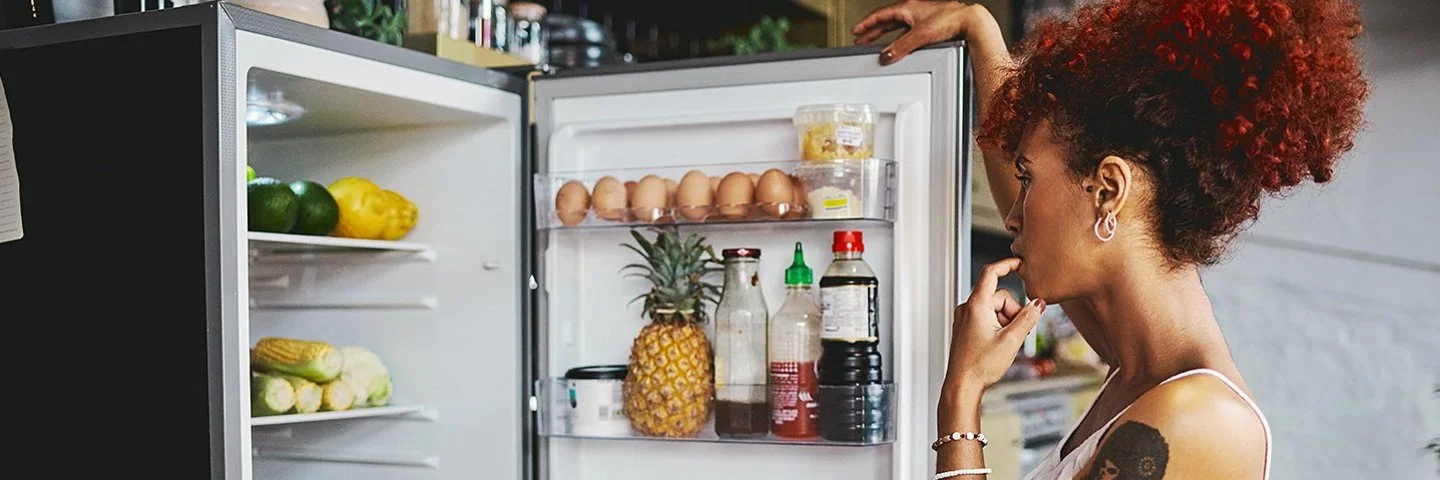 Eine Frau in einem sommerlichen Outfit steht überlegend vor dem gut gefüllten Kühlschrank, die geöffnetet Kühlschranktür in der Hand.