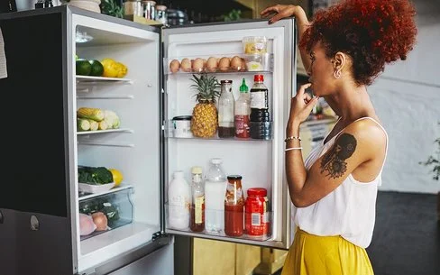 Eine Frau in einem sommerlichen Outfit steht überlegend vor dem gut gefüllten Kühlschrank, die geöffnetet Kühlschranktür in der Hand.