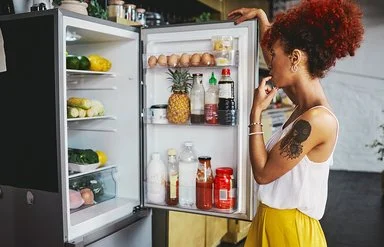 Eine Frau in einem sommerlichen Outfit steht überlegend vor dem gut gefüllten Kühlschrank, die geöffnetet Kühlschranktür in der Hand.
