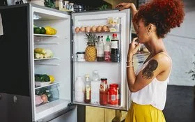 Eine Frau in einem sommerlichen Outfit steht überlegend vor dem gut gefüllten Kühlschrank, die geöffnetet Kühlschranktür in der Hand.