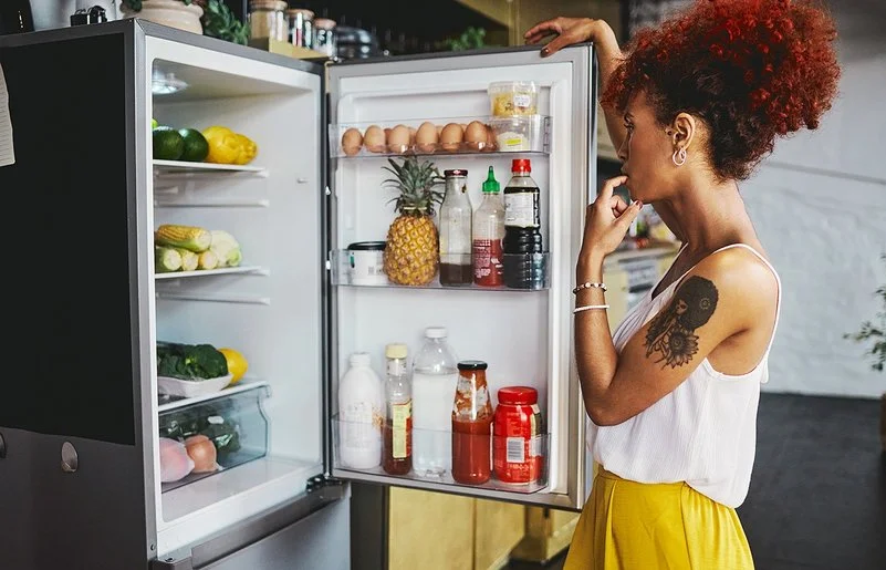 Eine Frau in einem sommerlichen Outfit steht überlegend vor dem gut gefüllten Kühlschrank, die geöffnetet Kühlschranktür in der Hand.