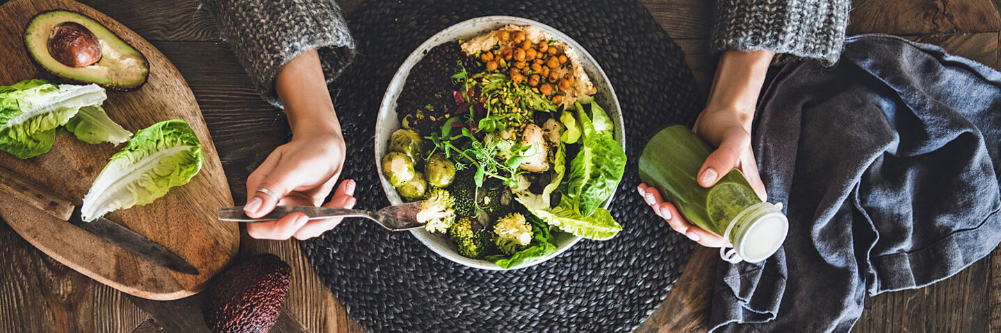 Person isst eine selbstgemachte Bowl mit frischem Salat, Avocado und Kichererbsen und hält eine Flasche mit grünem Smoothie in der linken Hand.