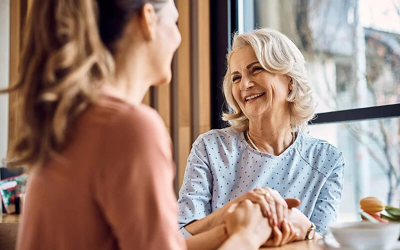 Eine erwachsene Tochter und ihre Mutter sitzen sich händehaltend in einem Café gegenüber, lächeln und genießen ihr Gespräch.
