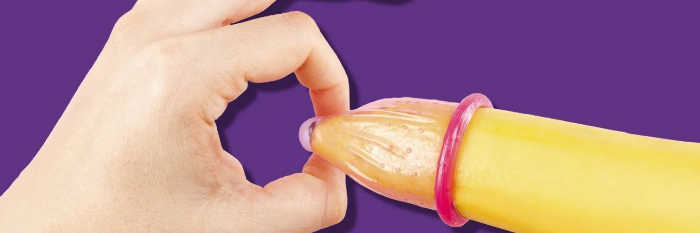 Hand mit roten Fingernägeln hält das Ende eines Kondoms auf einer Banane.