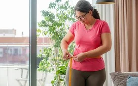 Eine Frau in Sportkleidung steht in einem Wohnraum und misst ihren Taillenumfang.