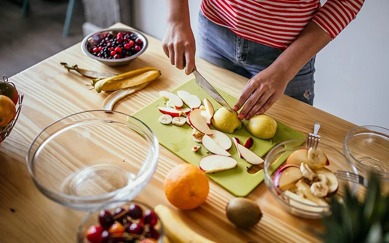 Eine Person schneidet auf einem Schneidebrett Birnen und Äpfel in Scheiben. Auf dem Tisch liegen eine Banane, eine Orange, eine Schale mit Beeren, eine Schale mit Bananenscheiben und zwei leere Glasschüsseln.