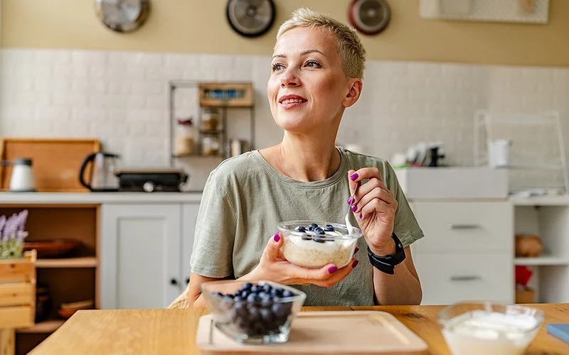 Frau sitzt in der Küche. Sie hält eine Schüssel mit Joghurt und frischen Blaubeeren in der Hand und genießt die gesunde Mahlzeit.
