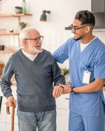 Ein männlicher Pfleger stützt einen älteren Mann mit Gehhilfe in seiner Küche.
