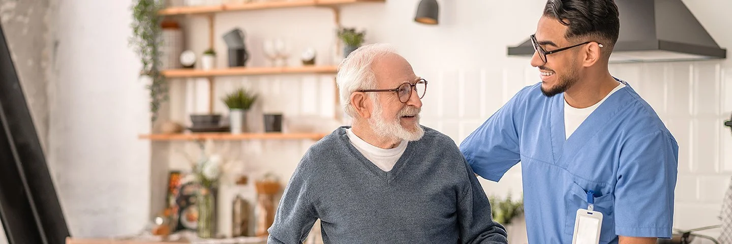 Ein männlicher Pfleger stützt einen älteren Mann mit Gehhilfe in seiner Küche.