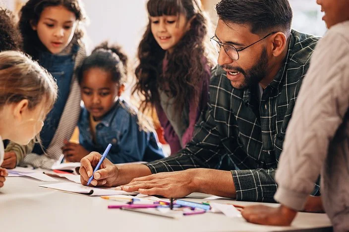 Ein Erzieher oder Grundschullehrer ist von vielen Kindern umgeben. Er schreibt etwas auf.