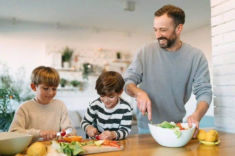 Ein Vater und zwei Jungen schneiden Gemüse und bereiten gemeinsam in einer hellen Küche einen Salat zu