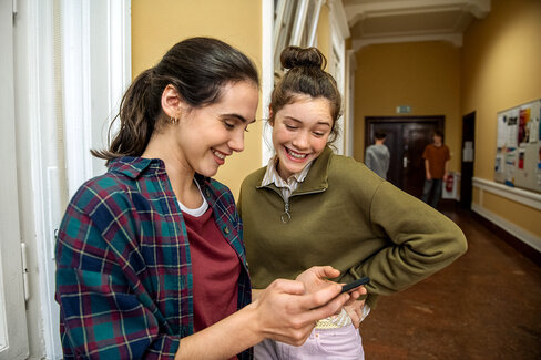 Zwei Schülerinnen informieren sich auf einem Smartphone zum Thema Suchtprävention.