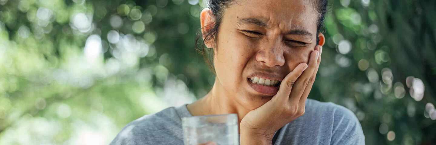Eine junge Frau fasst sich an die Wange und hat ein schmerzverzerrtes Gesicht nach einem Schluck heißen Kaffees.