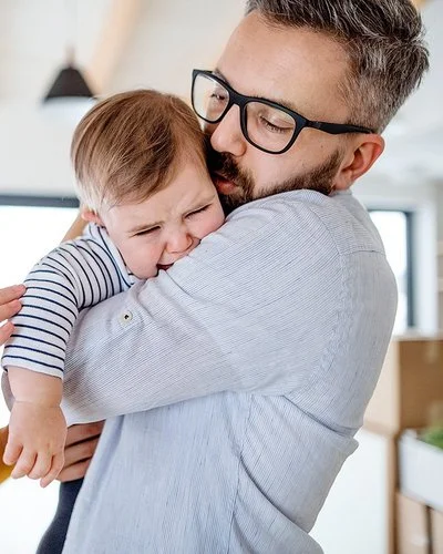 Weinendes Kleinkind auf dem Arm des Vaters, beide Eltern versuchen, das Kind zu beruhigen.