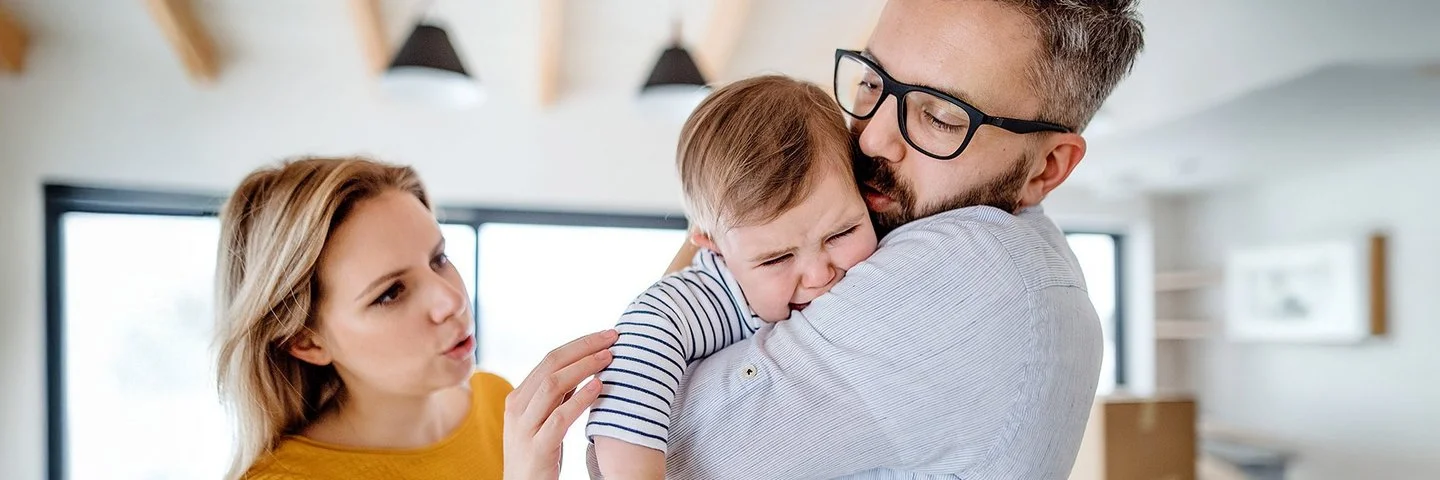 Weinendes Kleinkind auf dem Arm des Vaters, beide Eltern versuchen, das Kind zu beruhigen.