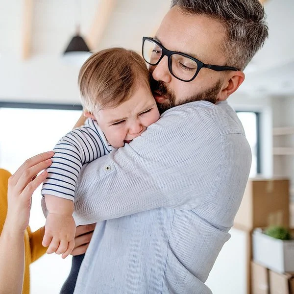 Weinendes Kleinkind auf dem Arm des Vaters, beide Eltern versuchen, das Kind zu beruhigen.