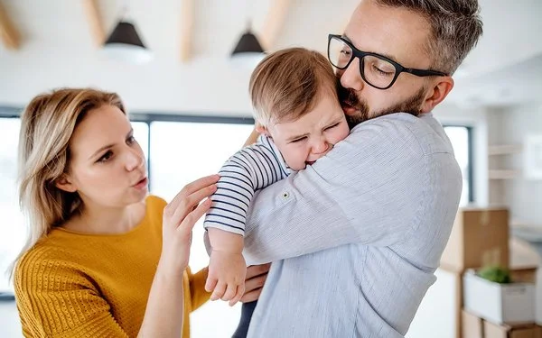 Weinendes Kleinkind auf dem Arm des Vaters, beide Eltern versuchen, das Kind zu beruhigen.