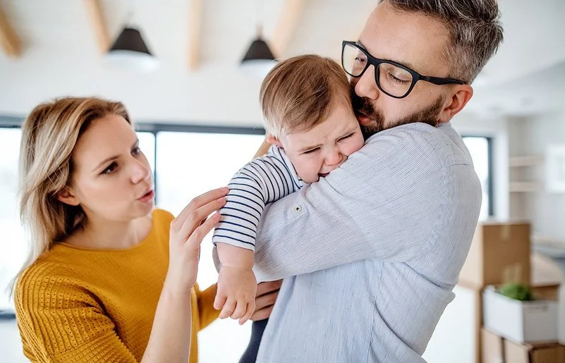Weinendes Kleinkind auf dem Arm des Vaters, beide Eltern versuchen, das Kind zu beruhigen.