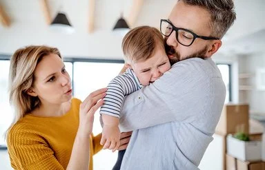 Weinendes Kleinkind auf dem Arm des Vaters, beide Eltern versuchen, das Kind zu beruhigen.