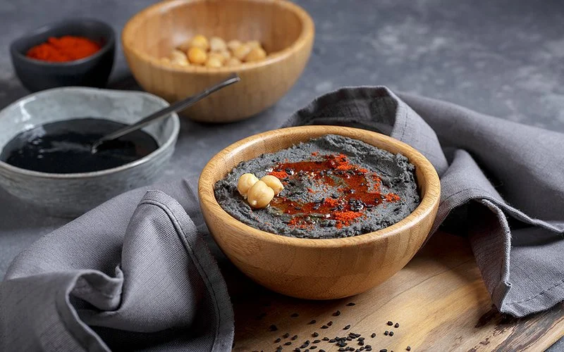 Schwarzer Hummus in einer kleinen Schüssel auf einem Holzbrett, im Hintergrund kleine Schüsseln mit Kicherebsen und schwarzem Sesam-Tahin.