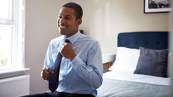 Ein junger Mann in hellblauem Hemd sitzt auf einem Bett und bindet sich eine Krawatte um den Hals.