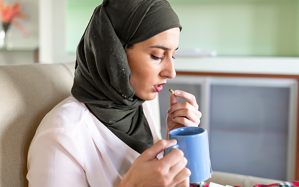 Eine junge Frau mit Hijab nimmt eine Kapsel ein und hält dabei einen blauen Becher in der anderen Hand.