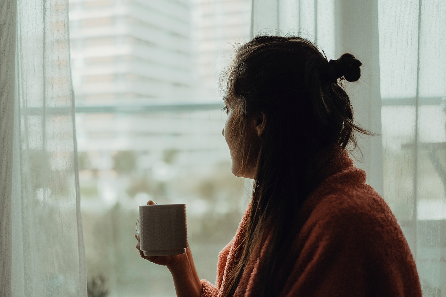 Frau mit Winterdepression schaut aus dem Fenster, sie hält dabei eine Tasse in der Hand.