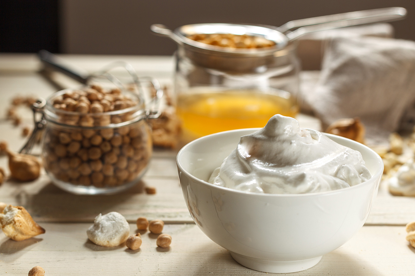 Auf einem Holztisch stehen ein Einmachglas Kichererbsen, eine Schüssel mit Abtropfwasser und aufgeschlagenes Aquafaba.