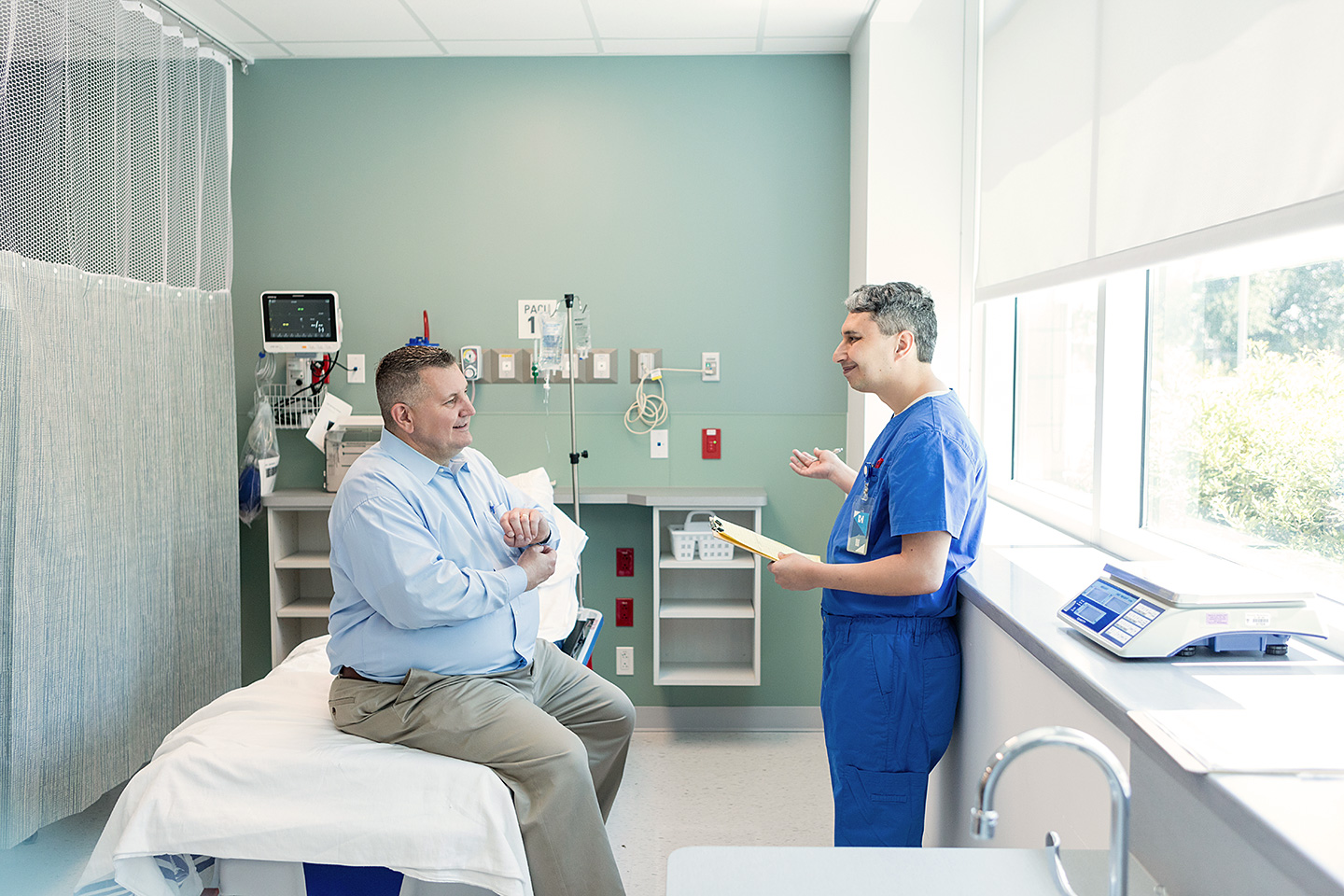 Ein von Übergewicht betroffener Mann sitzt auf einer Liege in einem Untersuchungsraum einer Klinik oder eines medizinischen Zentrums. Er unterhält sich mit einem Arzt, der ihm gegenübersteht.