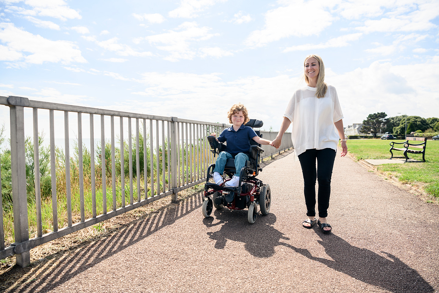 Mutter geht Hand in Hand mit ihrem Kind, das wegen einer Behinderung im Rollstuhl sitzt, spazieren.