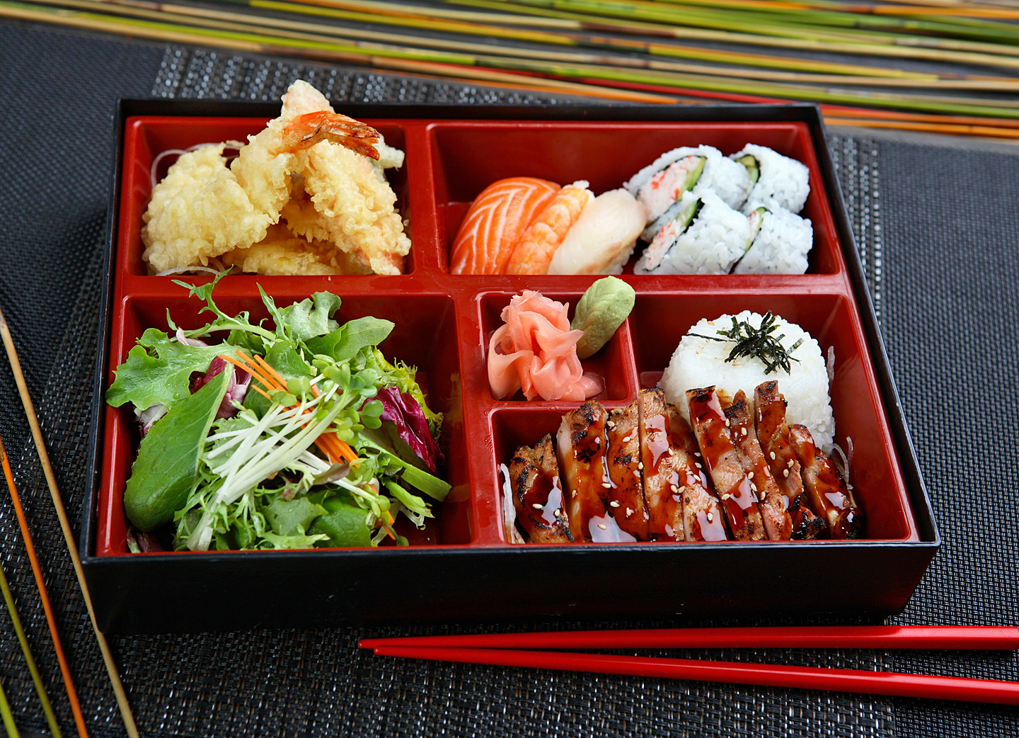 Klassische japanische Bento-Box mit 5 unterschiedlich großen Fächern mit Sushi, Teriyaki-Huhn, Tempura-Shrimps und Salat.