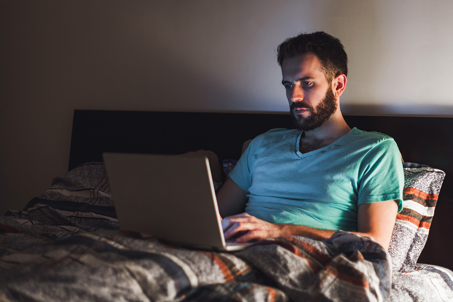 Ein Mann sitzt mit angeschaltetem Laptop im Bett, das davon ausgehende blaue Licht könnte seinen Schlaf negativ beeinträchtigen.