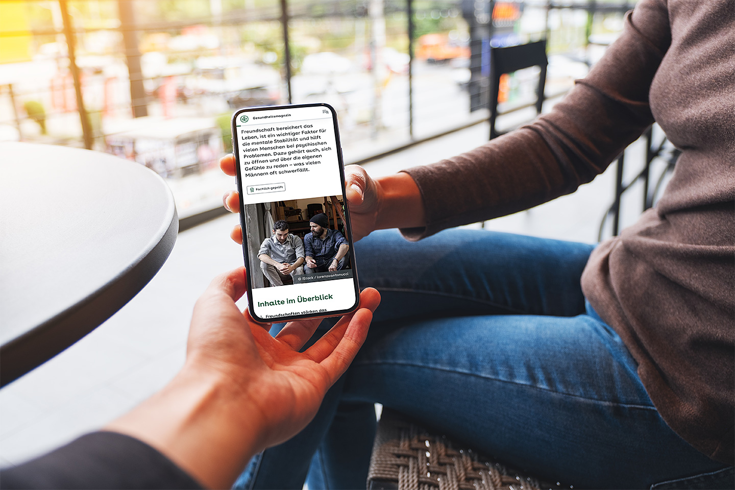 Eine Frau sitzt auf einer Terrasse und hält ein Smartphone in der Hand. Sie zeigt einer anderen Person den Bildschirm, auf dem ein Artikel aus dem AOK-Gesundheitsmagazin zu sehen ist.