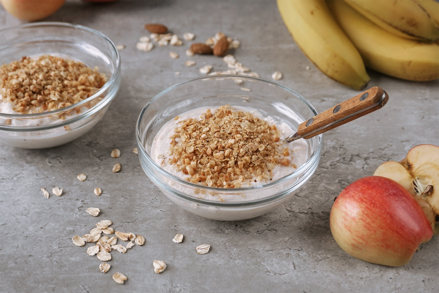 Auf grauem Hintergrund stehen zwei kleine Glasschalen, gefüllt mit Apfel-Zimt-Quark und Hafercrunch-Topping. Daneben sind zwei halbierte Äpfel arrangiert. Im Hintergrund verstreute Haferflocken und Mandeln sowie Bananen.