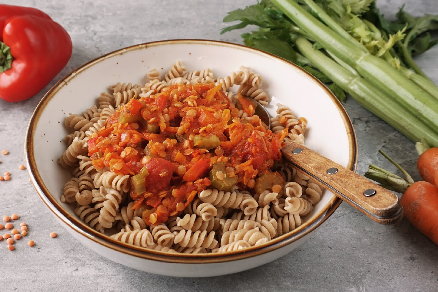 Weißer Teller mit Vollkorn-Fusilli und Gemüsebolognese, darin steckt ein Löffel mit Holzgriff. Daneben und dahinter liegen auf grauem Hintergrund einige rote Linsen, rote Paprika, Möhren und Stangensellerie.