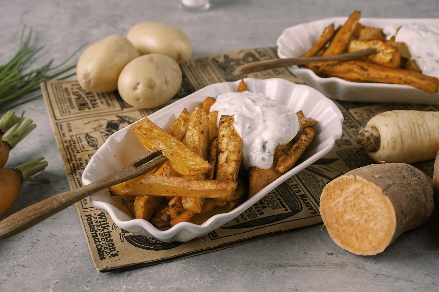 Weiße Schale mit Gemüsefritten, weißem Dip und Pommesgabel auf einem rustikalen Untergrund. Im Hintergrund Kartoffeln, Süßkartoffeln, Pastinaken und Möhren.