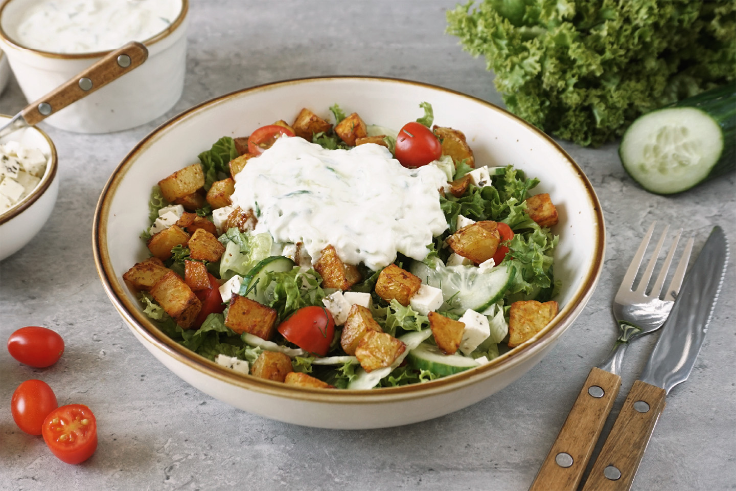 Weiße Schale mit knusprigen Kartoffelecken und selbstgemachtem Tsatsiki auf grünem Salat mit Fetawürfeln und Tomaten auf einem grauen Untergrund. Rechts daneben Messer und Gabel. Im Hintergrund Tomaten, Gurke und Lollo Rosso.