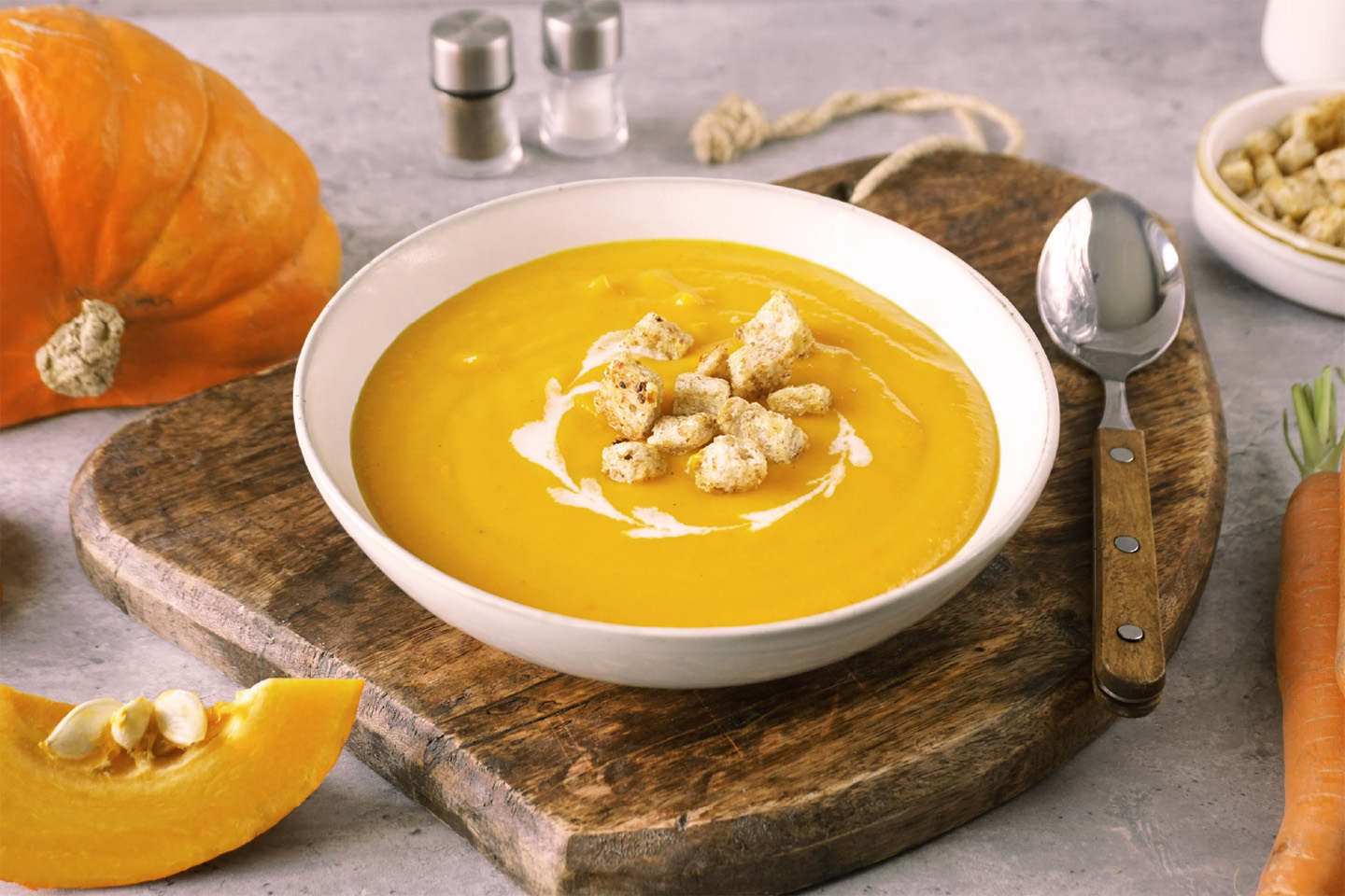 Weißer Teller mit cremiger, oranger Kürbissuppe und Croutons darauf. Der Untergrund besteht aus einem Holzbrett, rechts liegt ein Löffel. Im Hintergrund Möhren, Kürbis und eine weiße Schale mit Croutons.
