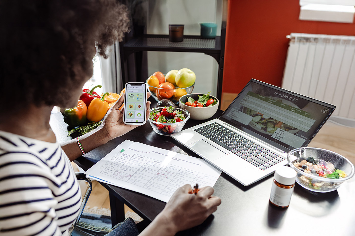 Eine Frau sitzt mit einem Smartphone am Tisch. Vor ihr liegen ein Ernährungsplan und gesunde Lebensmittel.