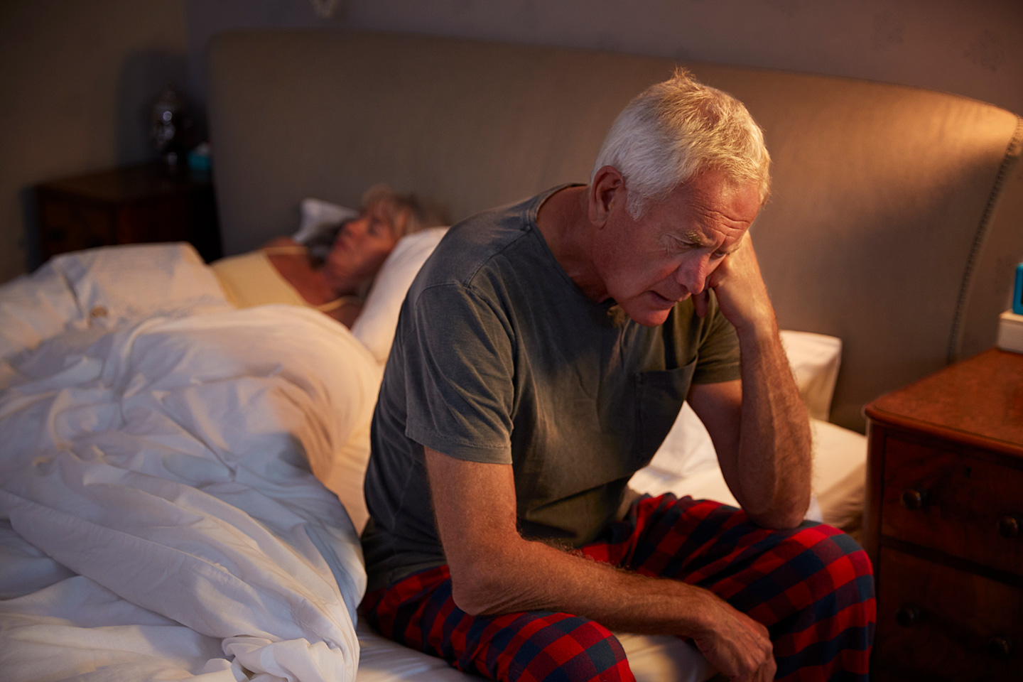Ein älterer Mann sitzt mit leicht gesenktem Kopf auf einem Bett und stützt den linken Arm ab.