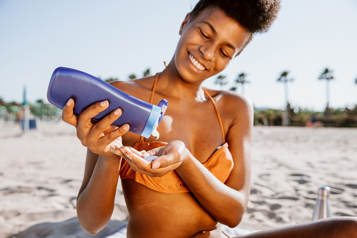 Eine junge Frau am Strand drückt Sonnencreme aus einer Flasche in die Hand, um sich vor der Sonne zu schützen.