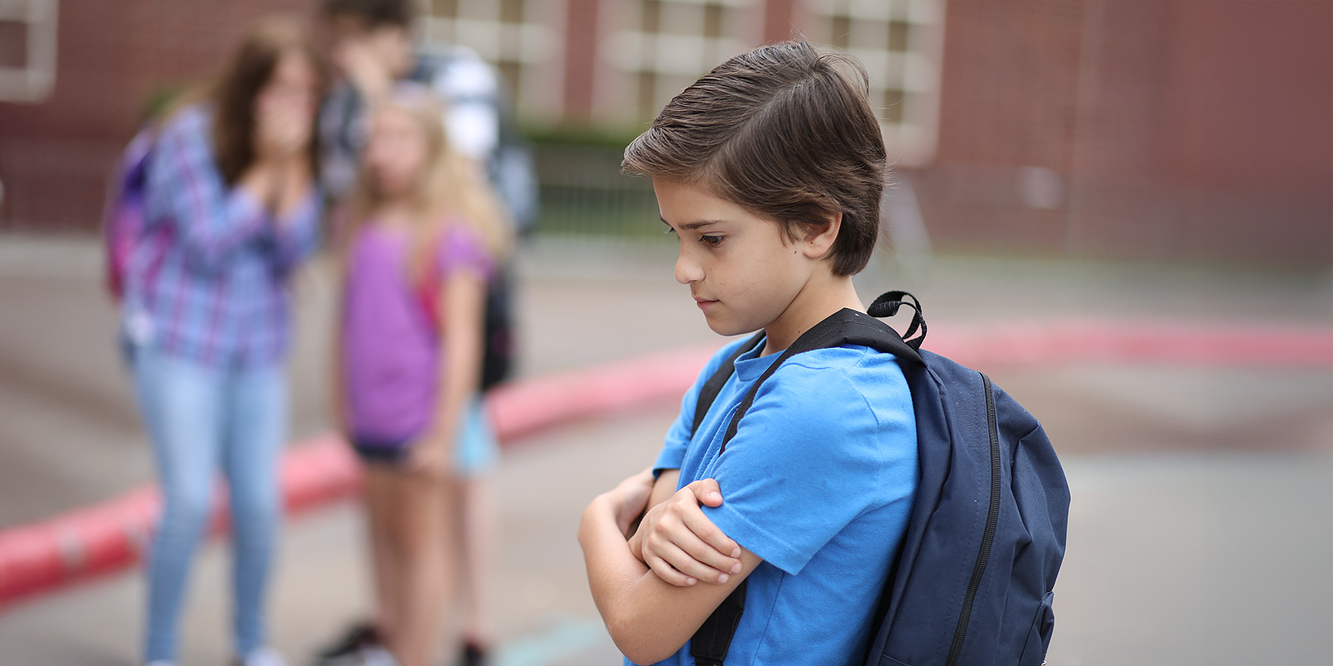 Ein Junge steht allein auf dem Schulhof und hält seine Arme vor die Brust. Im Hintergrund spielen Kinder.