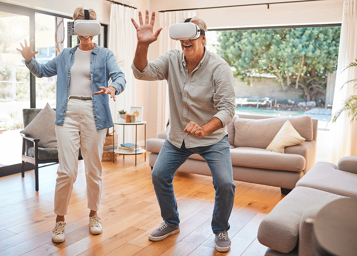 Zwei ältere Menschen mit einer VR-Brille bewegen sich im Wohnzimmer und lächeln.