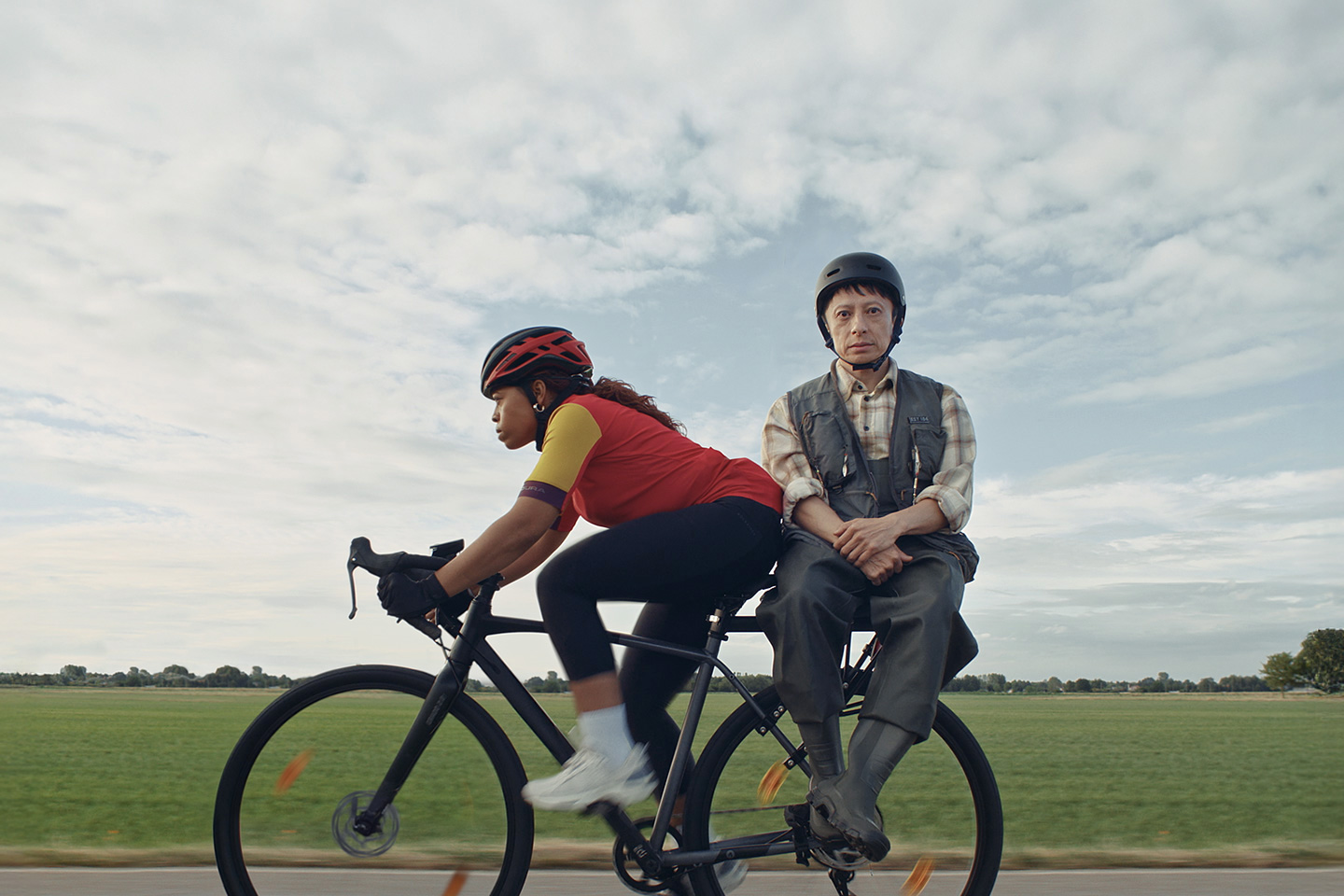 Zwei Menschen auf einem Fahrrad, das auf einer Landstraße an einem Feld vorbei fährt.