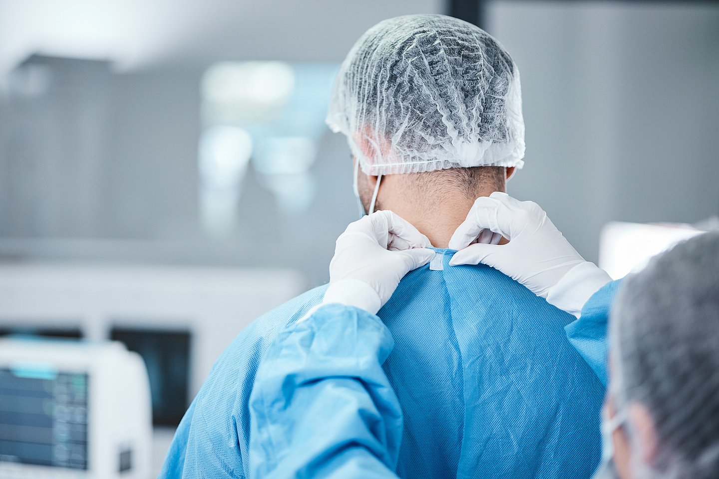 Ein Mediziner steht in einem OP und in OP-Kleidung, die Schutzkleidung wird auf der Rückseite von einer OP-Schwester mit behandschuhten Händen geschlossen.