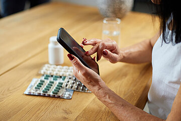 Eine Frau in Nahaufnahme sitzt am Tisch, auf dem verschiedene Tabletten liegen, und schaut auf ihr Handy.
